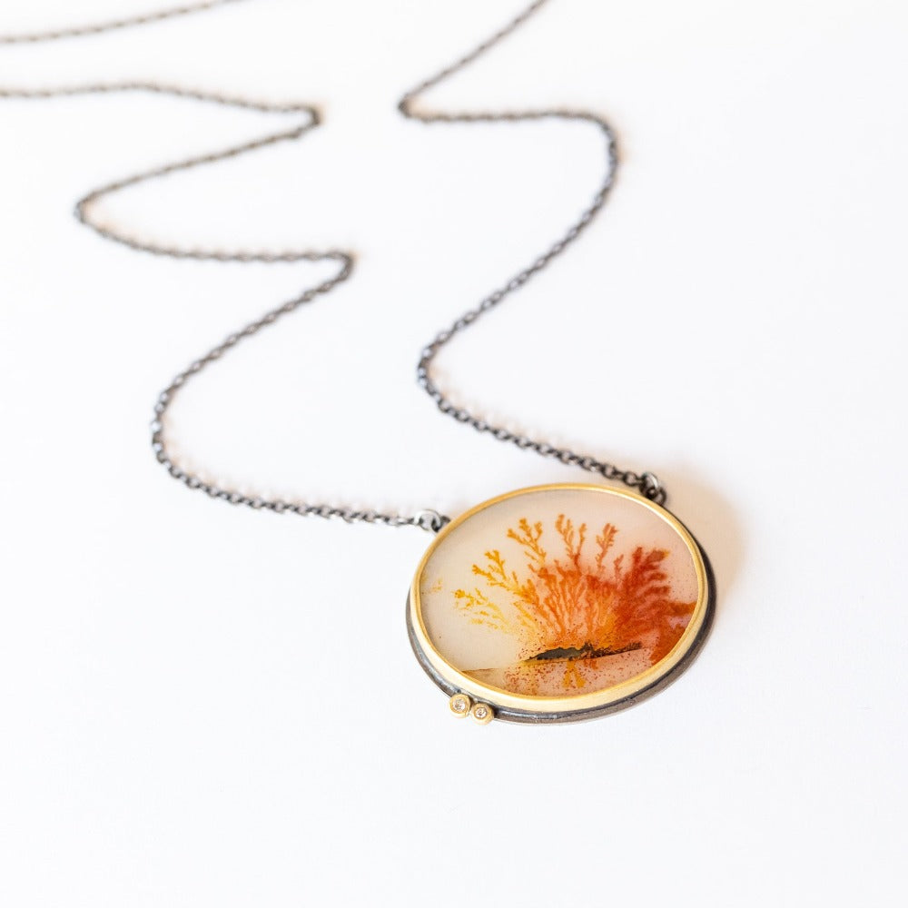 An oval shaped, orange dendritic agate is bezel set in yellow gold with two diamond accents. Necklace is sterling silver.