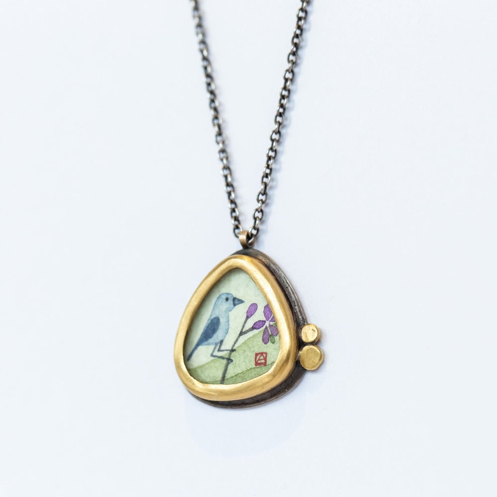 Ananda Khalsa pendant necklace featuring a hand painted bluebird on a branch, bezel set in yellow gold with tiny dot accents on a sterling silver chain.