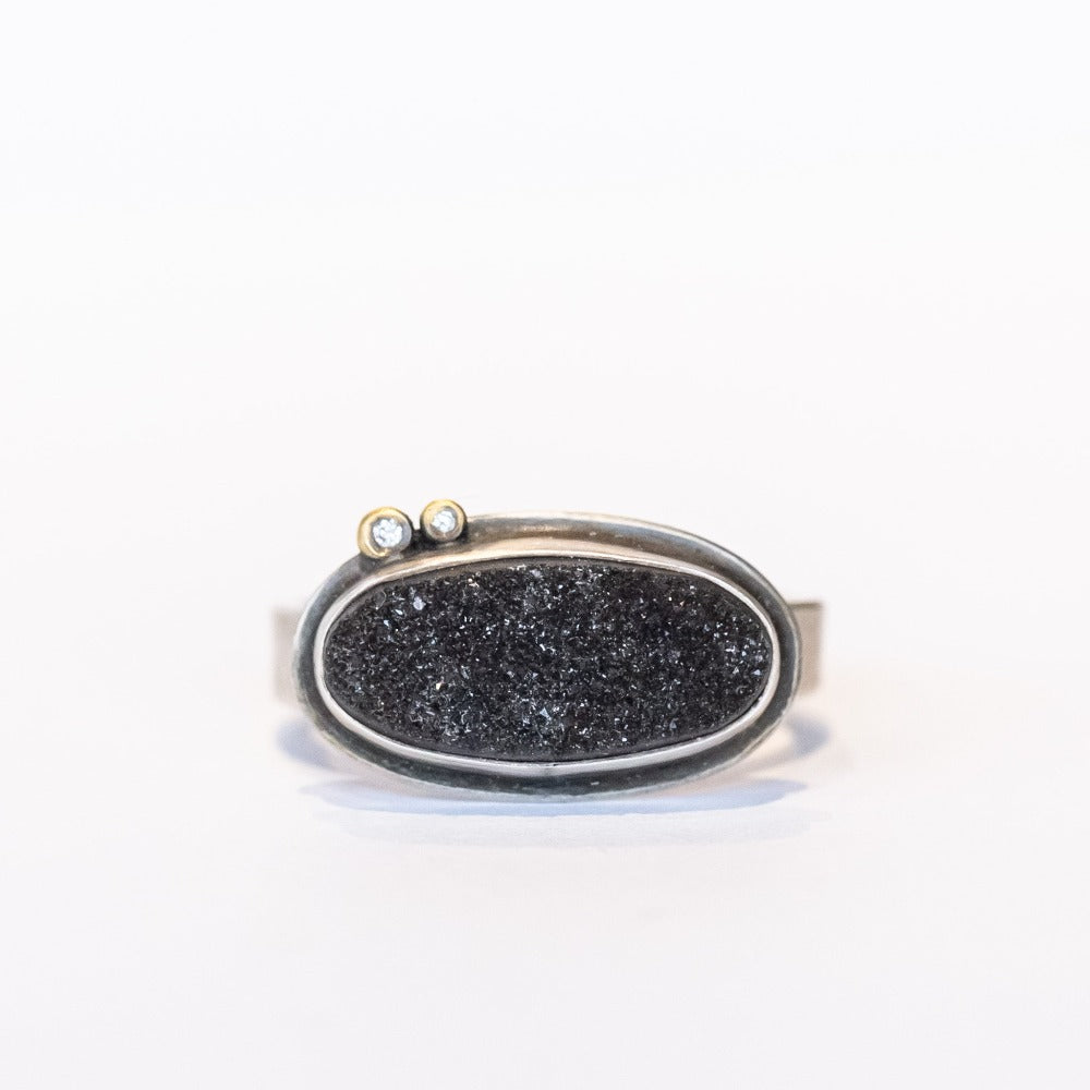 A horizontally set black oval drusy gemstone is bezel set on a sterling silver ring with two tiny diamond accents.
