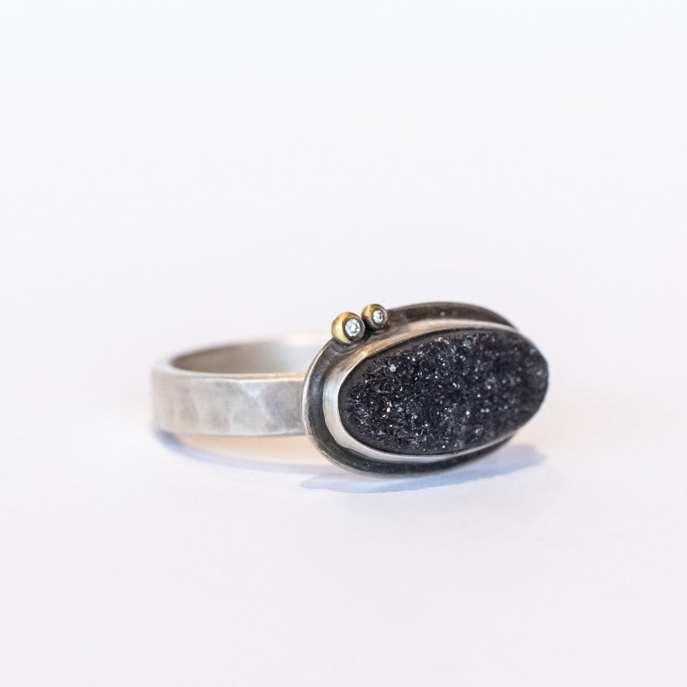 A horizontally set black oval drusy gemstone is bezel set on a sterling silver ring with two tiny diamond accents.