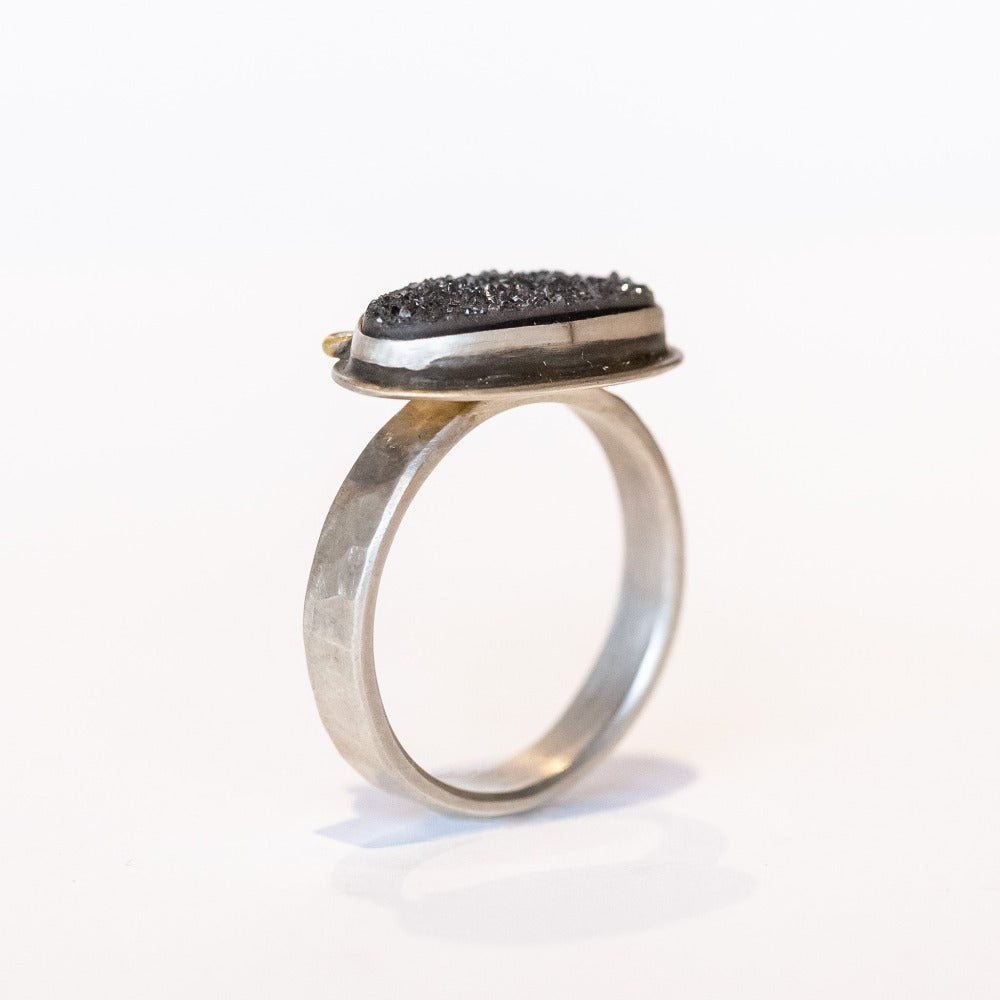 A horizontally set black oval drusy gemstone is bezel set on a sterling silver ring with two tiny diamond accents.