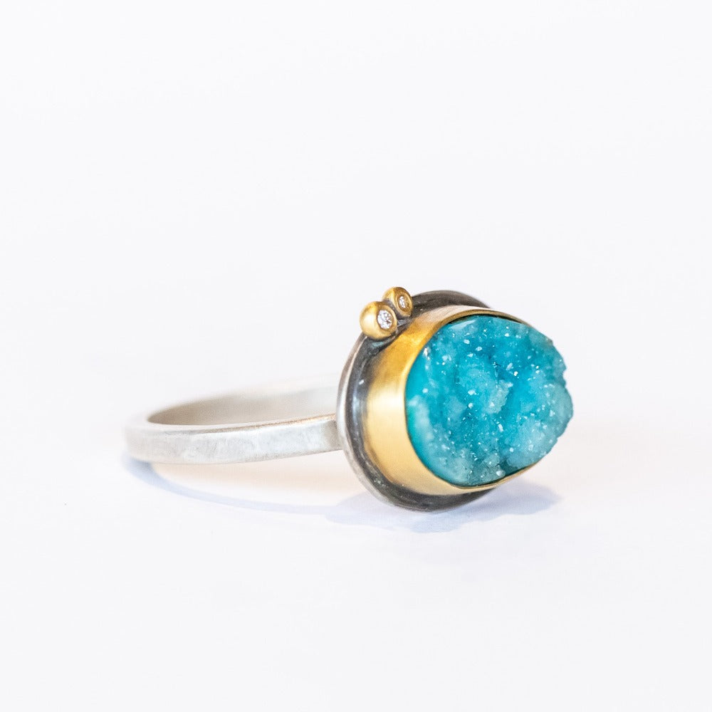 A bright, turquoise blue surface cut chrysocolla drusy ring set in a yellow gold bezel with two tiny diamond accents, on a sterling silver ring.