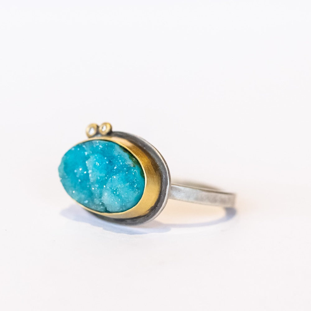 A bright, turquoise blue surface cut chrysocolla drusy ring set in a yellow gold bezel with two tiny diamond accents, on a sterling silver ring.