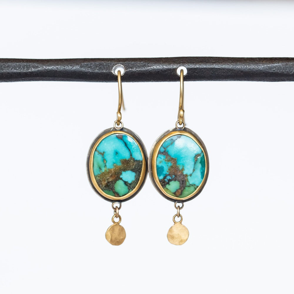 Drop earrings with bright green-blue oval turquoise gemstones, the stones have noticeable brown matrix veining, set in yellow gold with sterling silver backings. Small gold hammered dot accents dangle from the bottom. Ear hooks are gold.