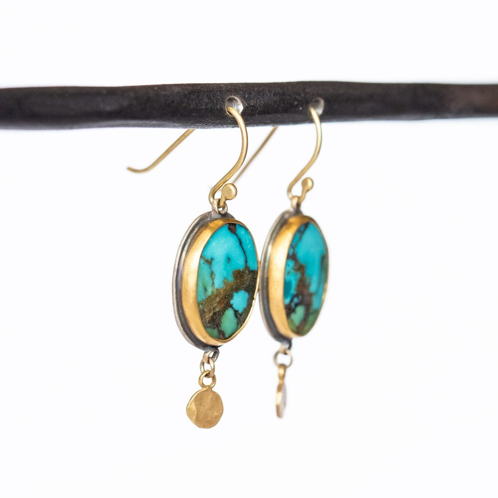 Drop earrings with bright green-blue oval turquoise gemstones, the stones have noticeable brown matrix veining, set in yellow gold with sterling silver backings. Small gold hammered dot accents dangle from the bottom. Ear hooks are gold.