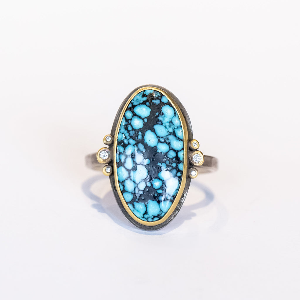 An elongated smooth oval matrix turquoise gemstone is bezel set in yellow gold, flanked by six tiny bezel set diamonds on a sterling silver ring.