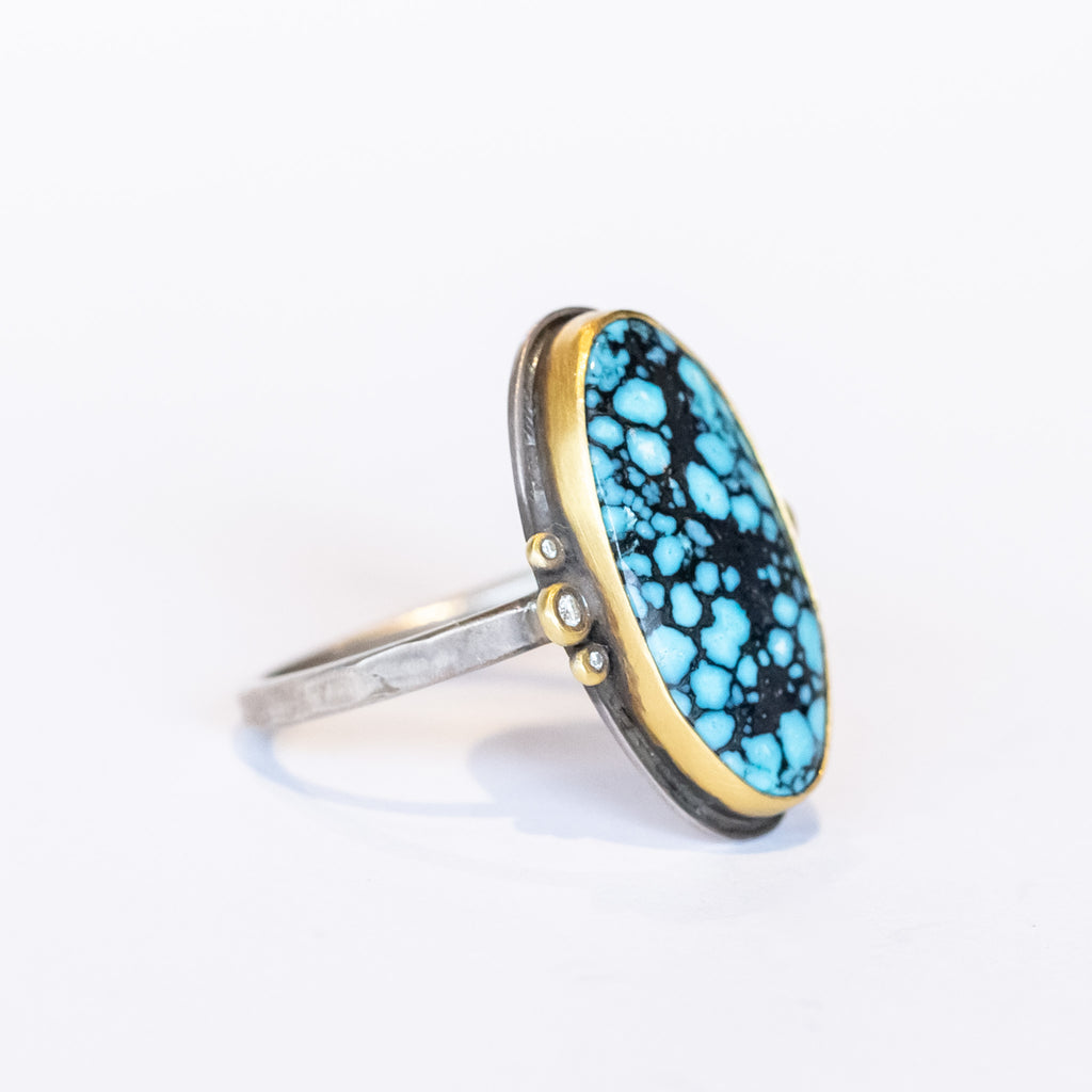 An elongated smooth oval matrix turquoise gemstone is bezel set in yellow gold, flanked by six tiny bezel set diamonds on a sterling silver ring.