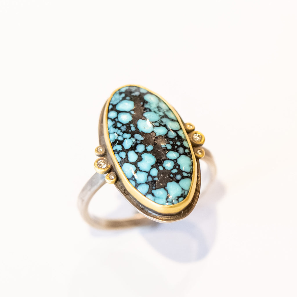 An elongated smooth oval matrix turquoise gemstone is bezel set in yellow gold, flanked by six tiny bezel set diamonds on a sterling silver ring.