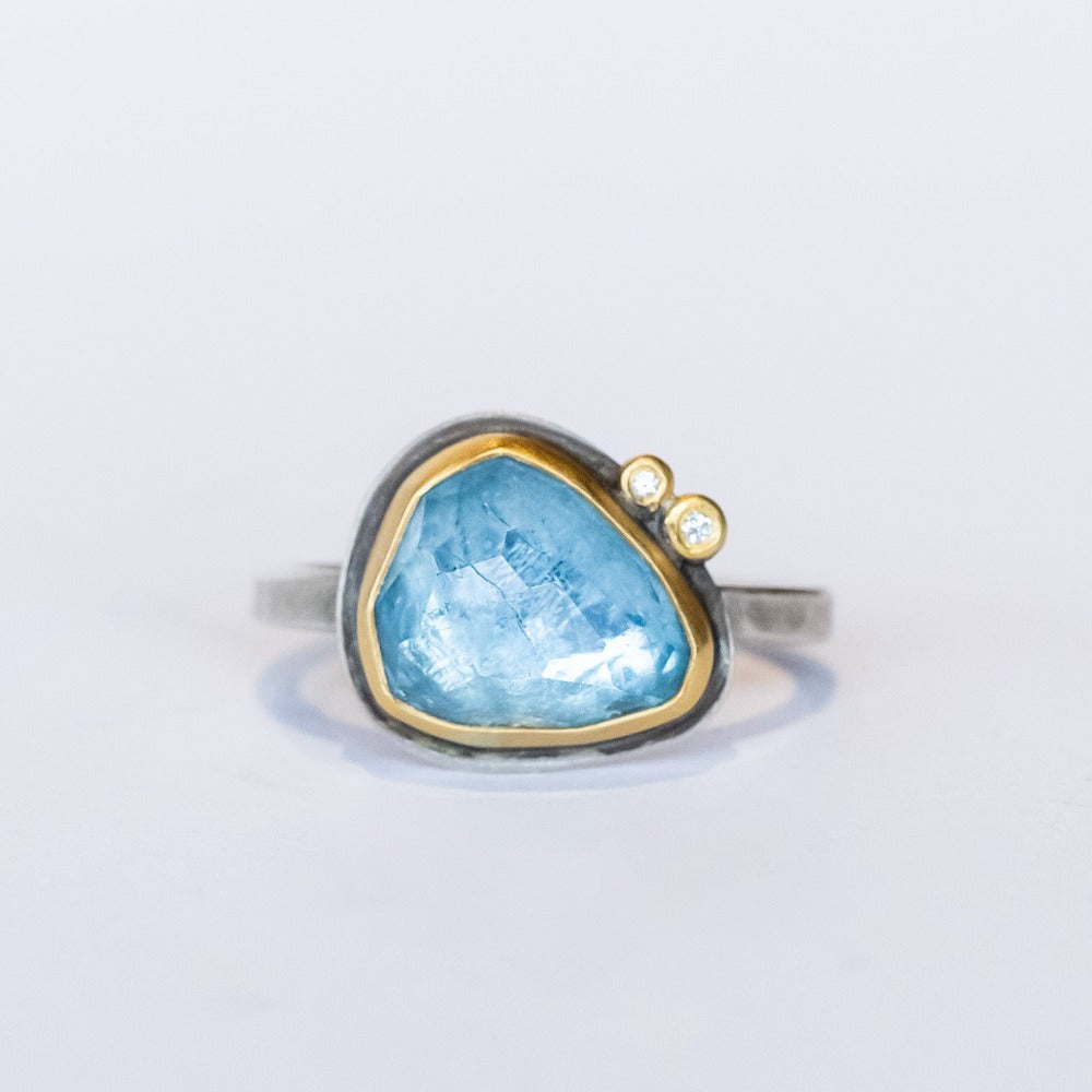 An asymmetrical triangular faceted blue topaz gemstone ring. The gem is bezel set in yellow gold, with two tiny diamond dot accents, on a sterling silver ring.