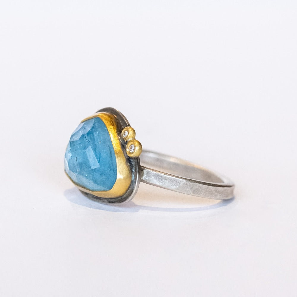 An asymmetrical triangular faceted blue topaz gemstone ring. The gem is bezel set in yellow gold, with two tiny diamond dot accents, on a sterling silver ring.