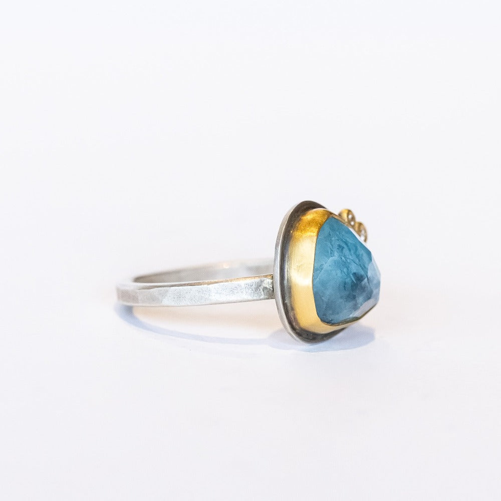 An asymmetrical triangular faceted blue topaz gemstone ring. The gem is bezel set in yellow gold, with two tiny diamond dot accents, on a sterling silver ring.