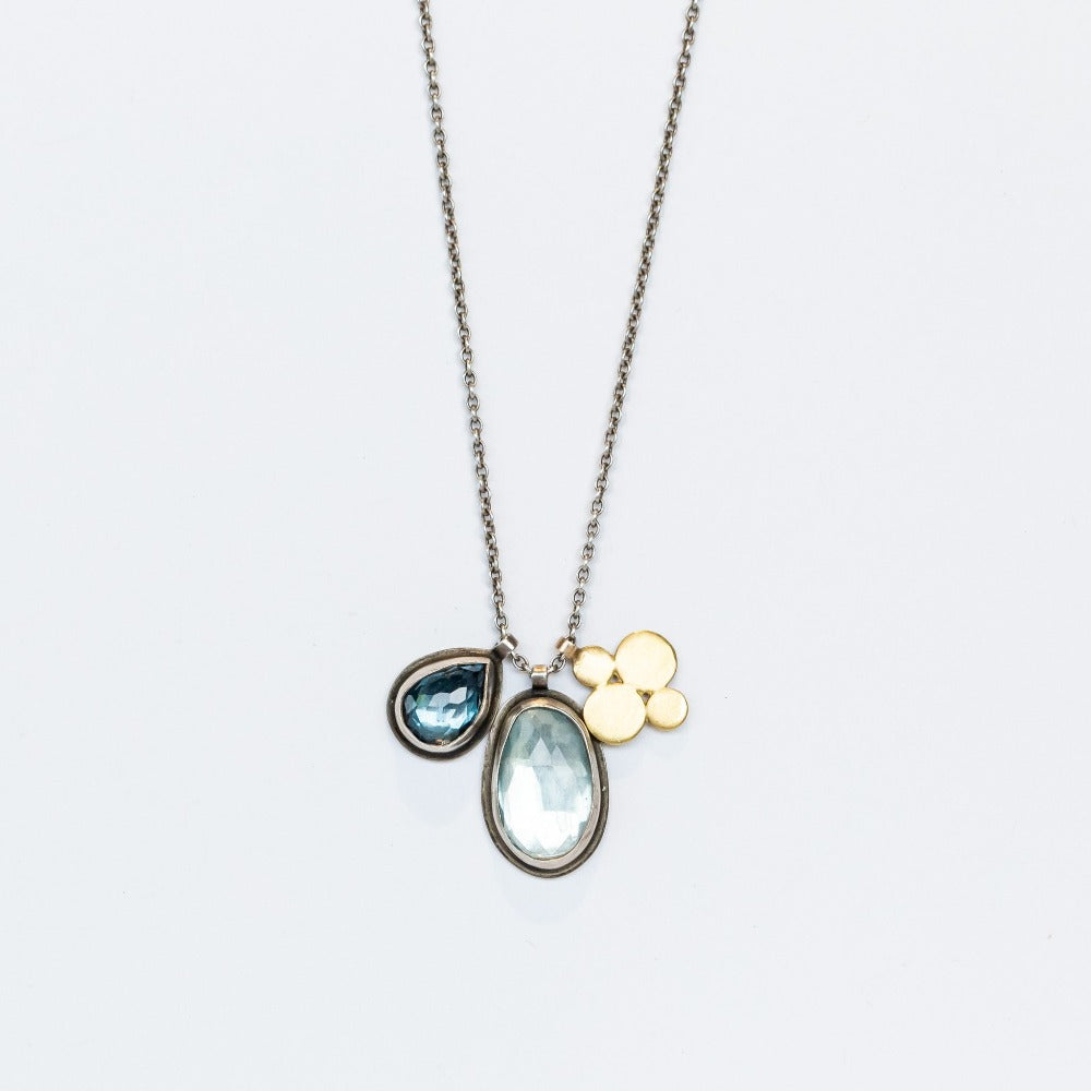 A sterling silver chain necklace with three charms: one bezel set teardrop london blue topaz, one bezel set oval blue topaz, and one gold flower shaped charm.