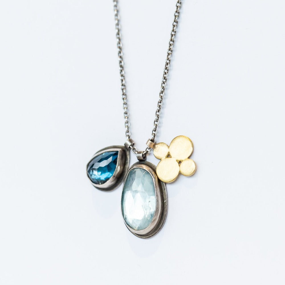 A sterling silver chain necklace with three charms: one bezel set teardrop london blue topaz, one bezel set oval blue topaz, and one gold flower shaped charm.