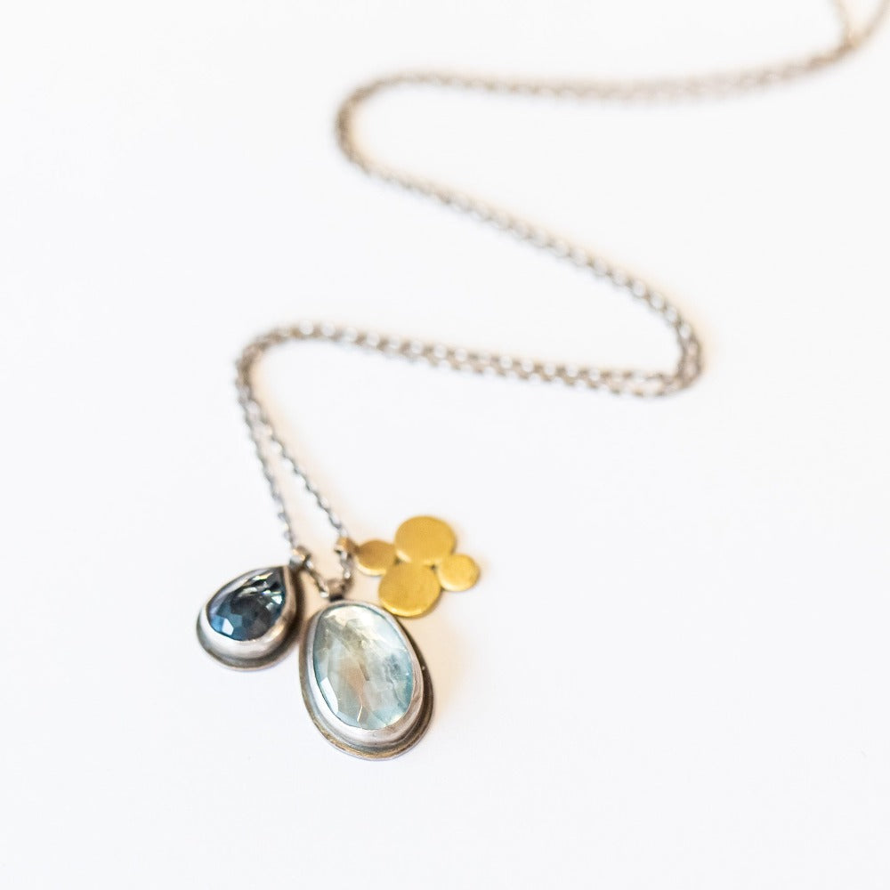 A sterling silver chain necklace with three charms: one bezel set teardrop london blue topaz, one bezel set oval blue topaz, and one gold flower shaped charm.