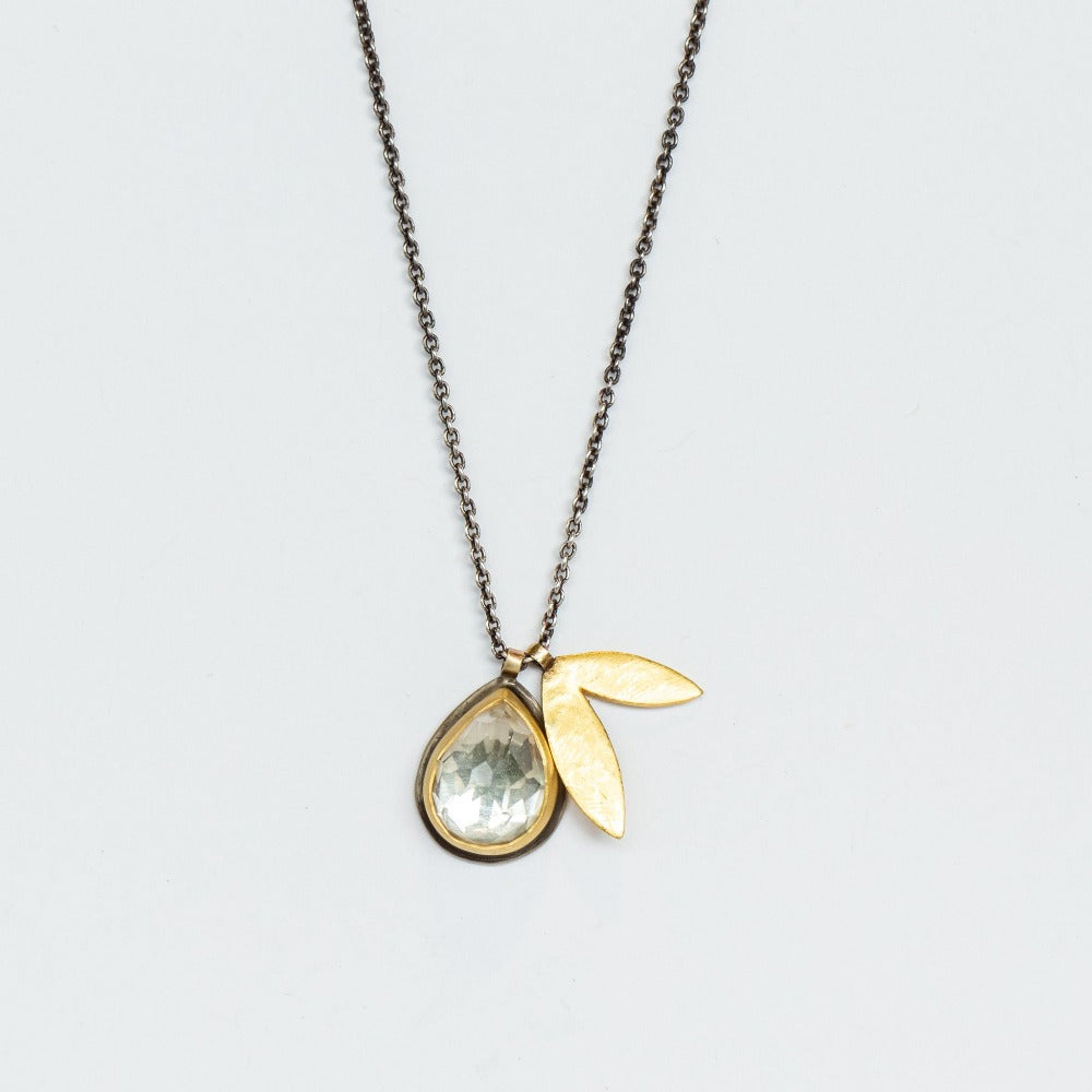 A clear, teardrop shaped topaz is bezel set in yellow gold with a silver backing, paired with a petal-shaped gold charm on an oxidized silver chain necklace.