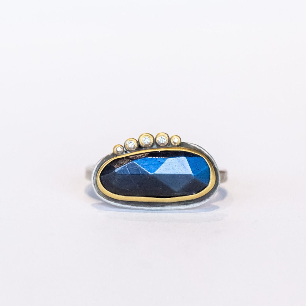 An elongated, rose cut labradorite gemstone in deep blue is set horizontally on this sterling silver ring. The gem is surrounded in a yellow gold bezel and accented along one edge with five petite diamonds.