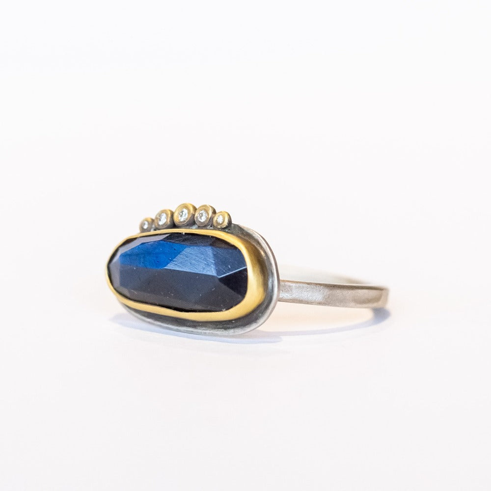 An elongated, rose cut labradorite gemstone in deep blue is set horizontally on this sterling silver ring. The gem is surrounded in a yellow gold bezel and accented along one edge with five petite diamonds.