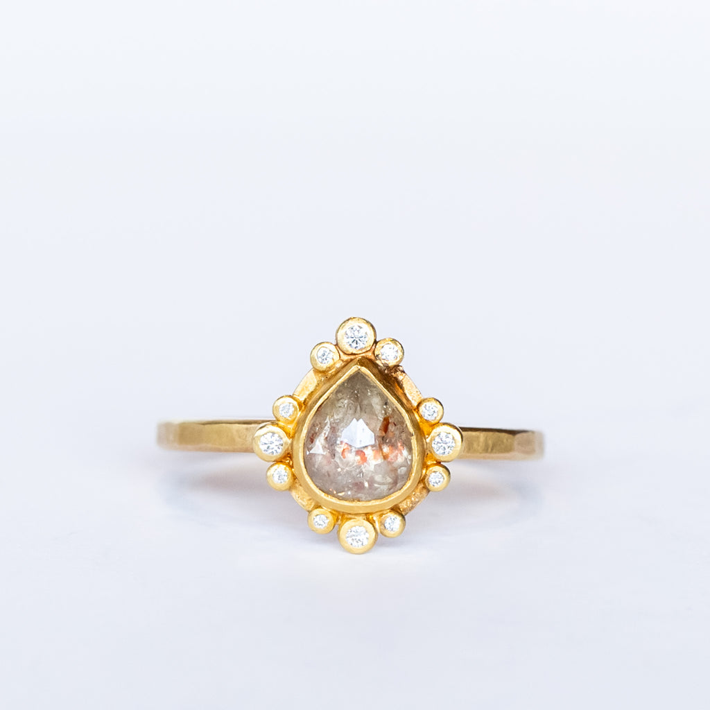 A teardrop shaped rose cut diamond with reddish inclusions is bezel set in yellow gold, surrounded by tiny round white diamonds in bezels on a yellow gold ring.
