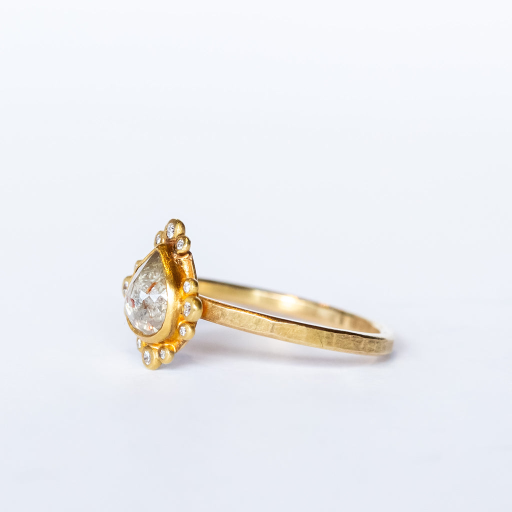 A teardrop shaped rose cut diamond with reddish inclusions is bezel set in yellow gold, surrounded by tiny round white diamonds in bezels on a yellow gold ring.