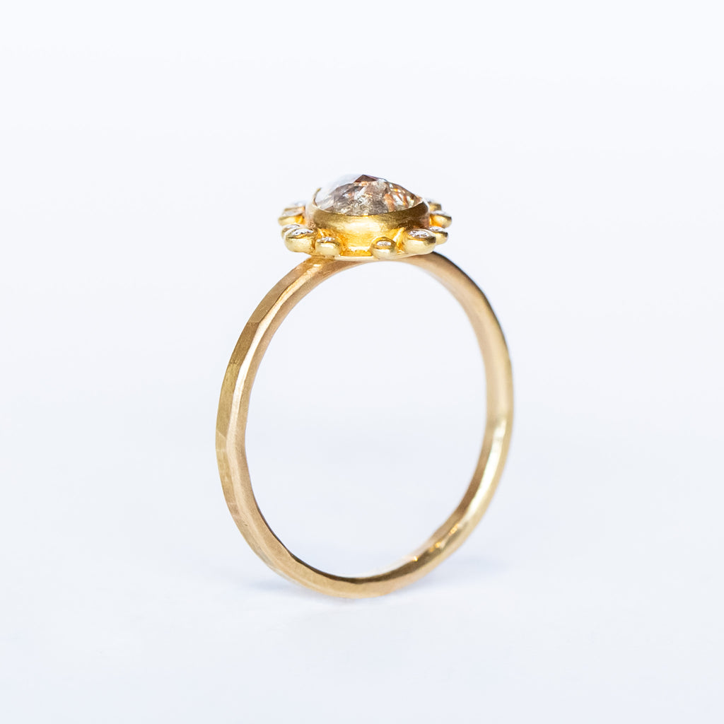 A teardrop shaped rose cut diamond with reddish inclusions is bezel set in yellow gold, surrounded by tiny round white diamonds in bezels on a yellow gold ring.