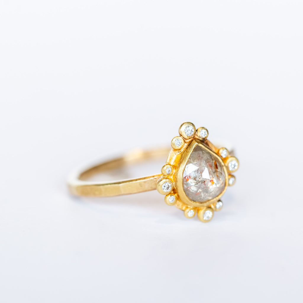 A teardrop shaped rose cut diamond with reddish inclusions is bezel set in yellow gold, surrounded by tiny round white diamonds in bezels on a yellow gold ring.