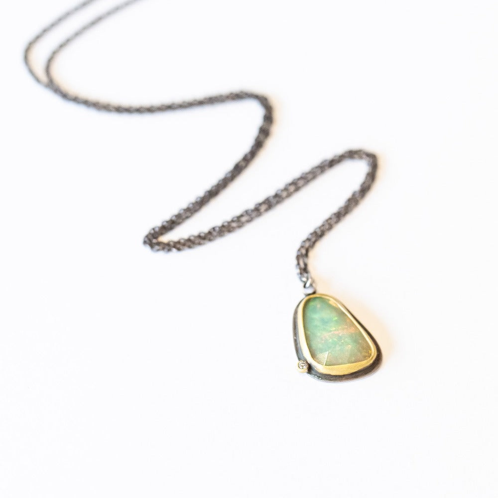An asymmetrical teardrop shaped opal pendant, bezel set in yellow gold backed by oxidized silver, with one tiny diamond accent on an oxidized silver necklace chain.