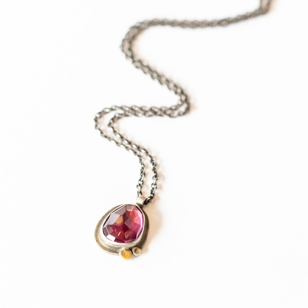 An asymmetrical teardrop shaped garnet is set in oxidized sterling silver with small gold dot accents.