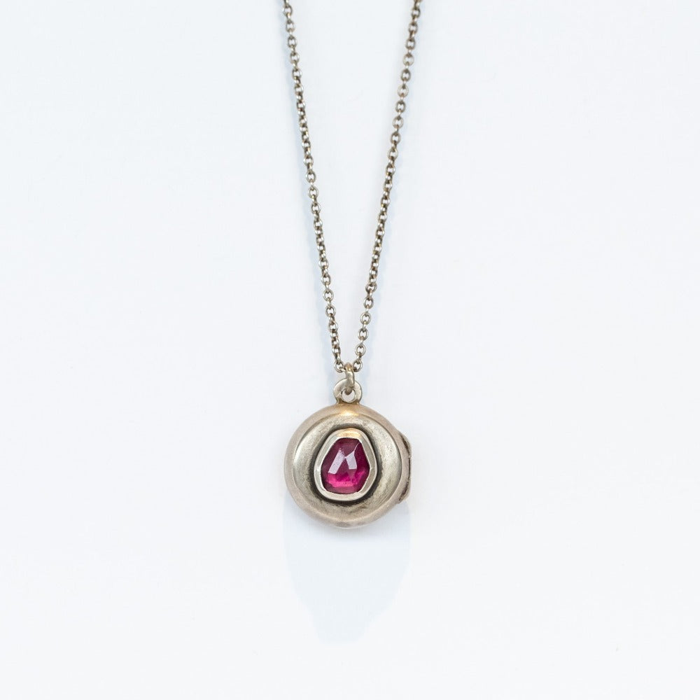 A handmade, round silver locket accented by an asymmetrical deep red garnet bezel set into its surface.