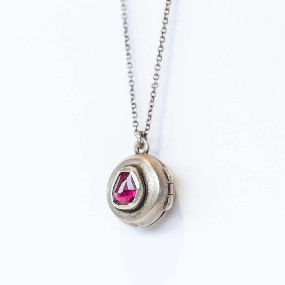 A handmade, round silver locket accented by an asymmetrical deep red garnet bezel set into its surface.