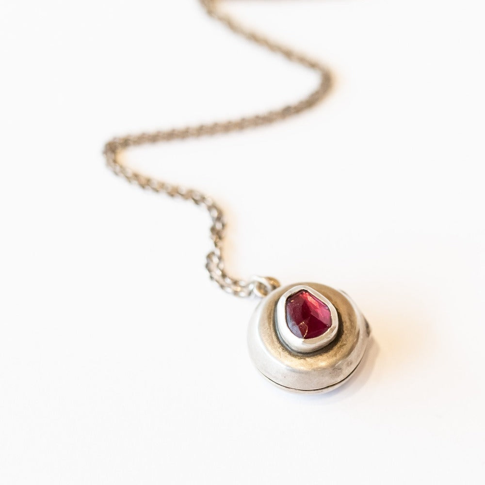 A handmade, round silver locket accented by an asymmetrical deep red garnet bezel set into its surface.
