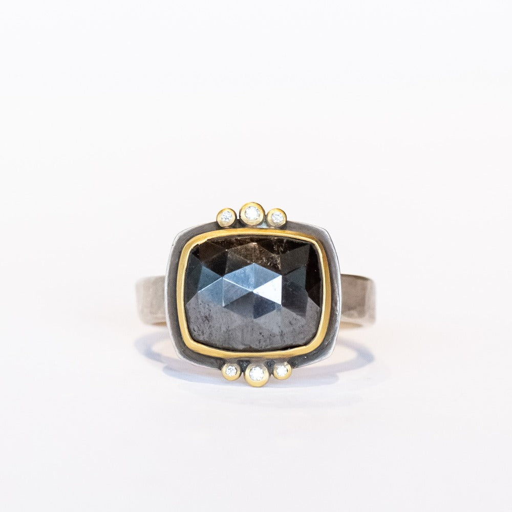 A rose cut, rectangular hematite gemstone is bezel set in yellow gold. It sits horizontally on this wide silver band, flanked on top and bottom by 3 tiny bezel set diamonds.