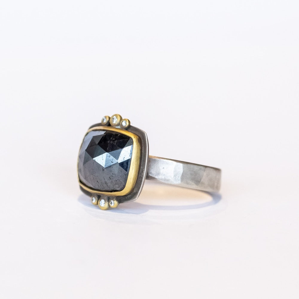 A rose cut, rectangular hematite gemstone is bezel set in yellow gold. It sits horizontally on this wide silver band, flanked on top and bottom by 3 tiny bezel set diamonds.