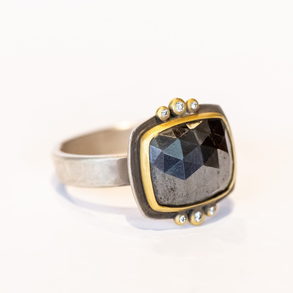 A rose cut, rectangular hematite gemstone is bezel set in yellow gold. It sits horizontally on this wide silver band, flanked on top and bottom by 3 tiny bezel set diamonds.