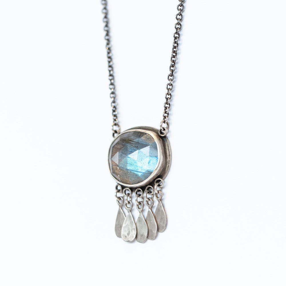 A large oval labradorite gemstone is bezel set in sterling silver with five hammered silver teardrop charms on this Ananda Khalsa necklace.