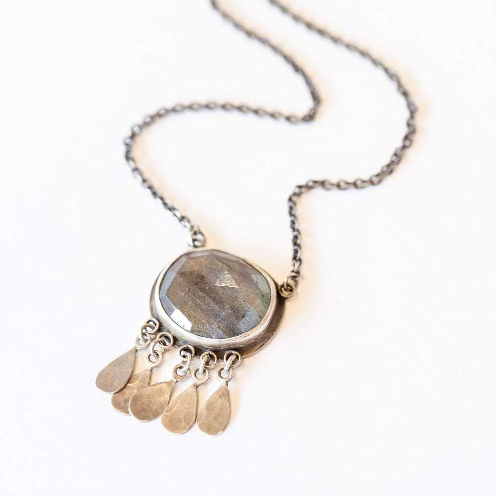 A large oval labradorite gemstone is bezel set in sterling silver with five hammered silver teardrop charms on this Ananda Khalsa necklace.