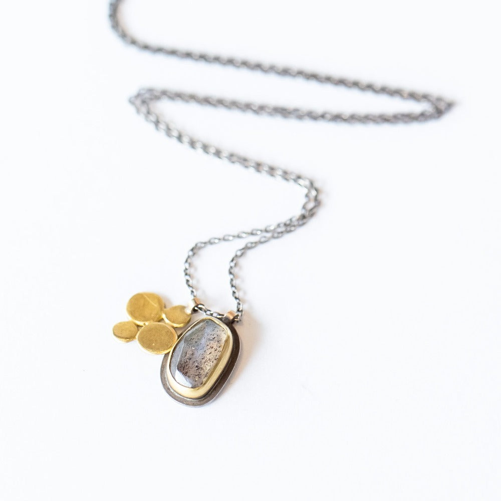 A gemstone pendant featuring a rose cut labradorite set in a 22 karat yellow gold bezel, backed by sterling silver, and accompanied by a 22 karat yellow gold disc charm detail. 