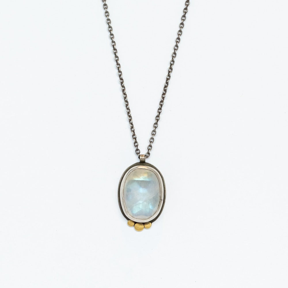 An oval, rose cut moonstone is bezel set in sterling silver with three dainty yellow gold dot accents underneath, suspended from an oxidized silver chain necklace.
