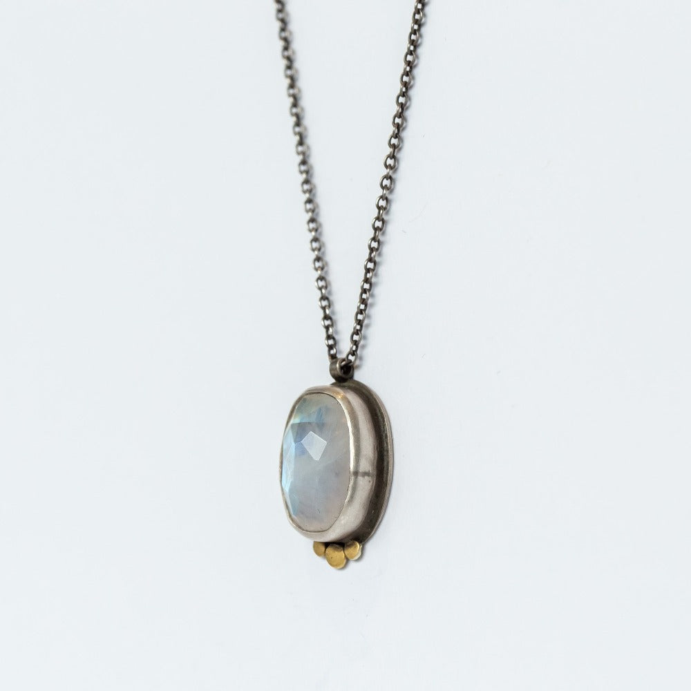 An oval, rose cut moonstone is bezel set in sterling silver with three dainty yellow gold dot accents underneath, suspended from an oxidized silver chain necklace.