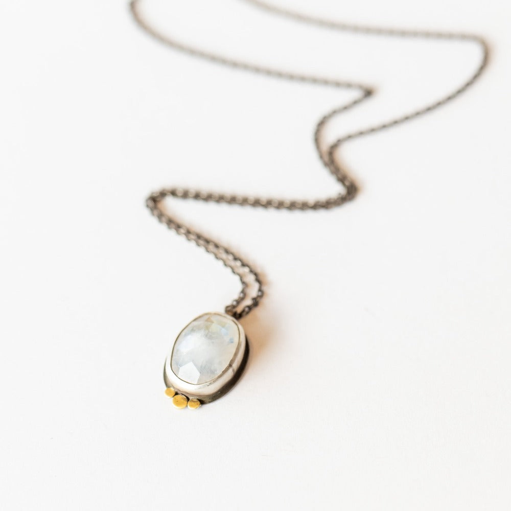An oval, rose cut moonstone is bezel set in sterling silver with three dainty yellow gold dot accents underneath, suspended from an oxidized silver chain necklace.