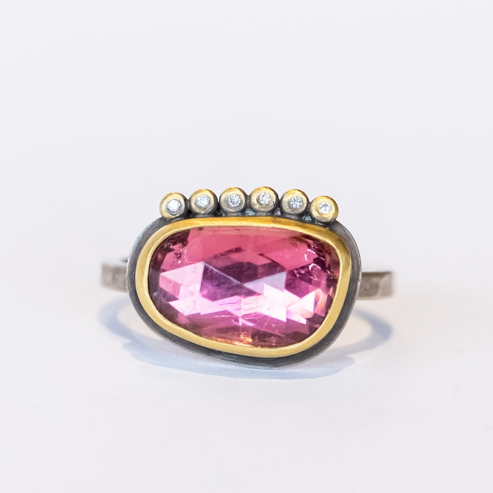 An elongated, asymmetrical pink tourmaline is bezel set in yellow gold with six tiny diamond accents on one side, all on a silver ring.