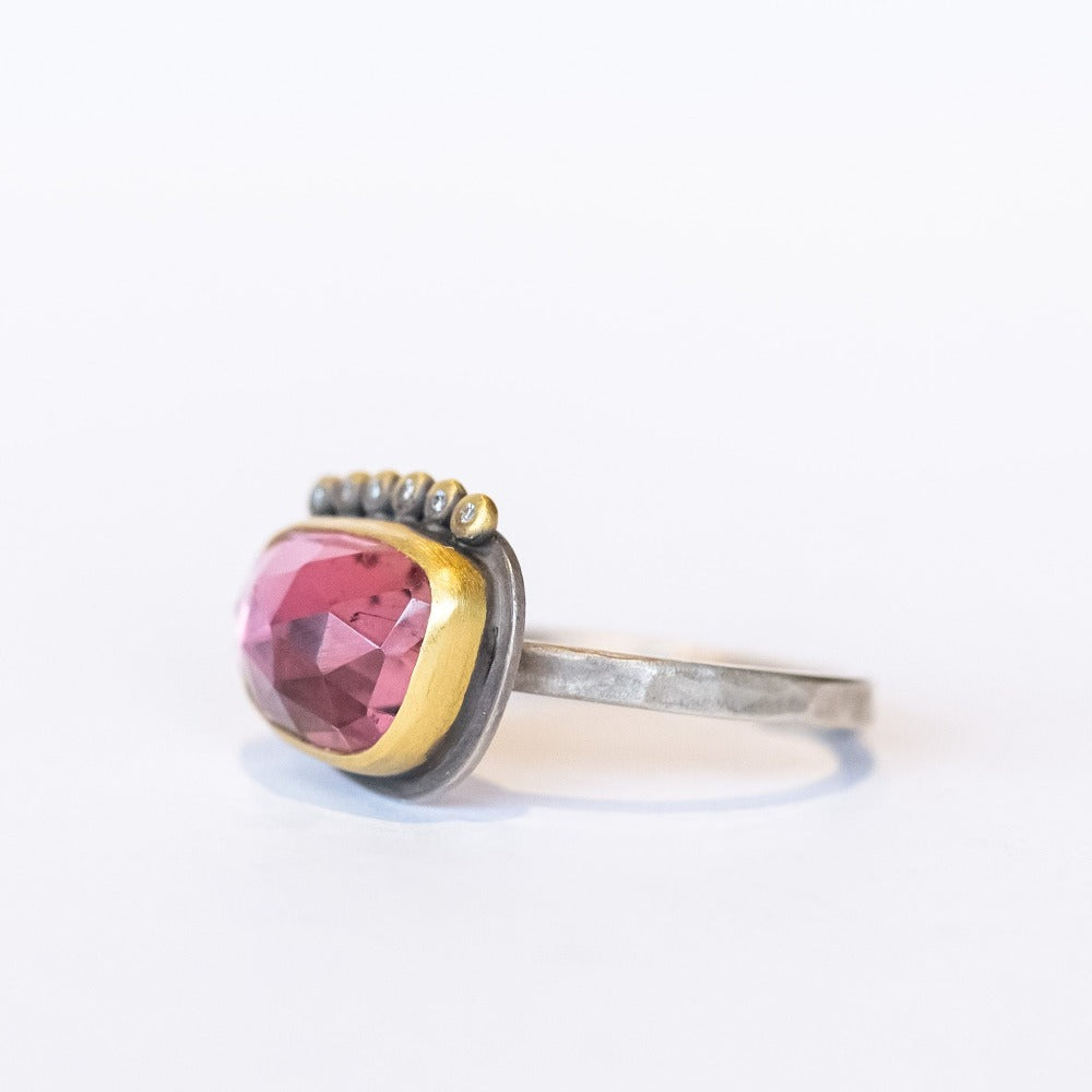 An elongated, asymmetrical pink tourmaline is bezel set in yellow gold with six tiny diamond accents on one side, all on a silver ring.