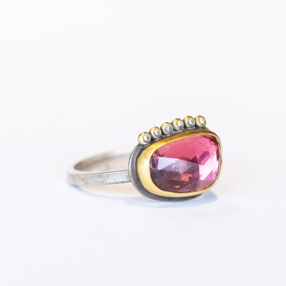 An elongated, asymmetrical pink tourmaline is bezel set in yellow gold with six tiny diamond accents on one side, all on a silver ring.