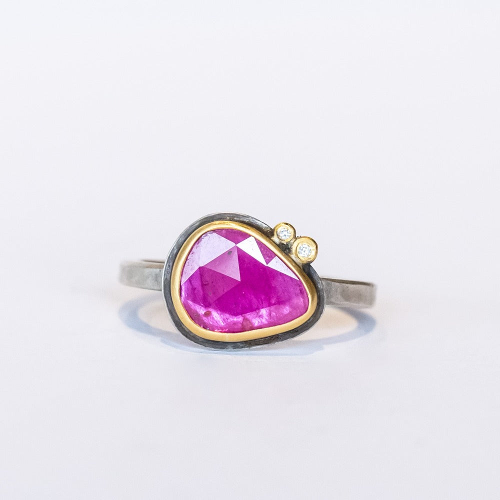 A horizontally set, teardrop shaped faceted pink ruby is bezel set in yellow gold mounted on a sterling silver ring. It is accented by two tiny bezel set diamonds.