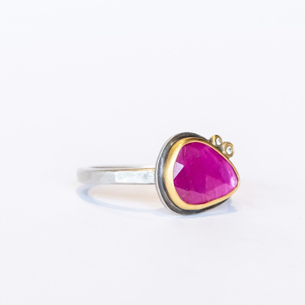 A horizontally set, teardrop shaped faceted pink ruby is bezel set in yellow gold mounted on a sterling silver ring. It is accented by two tiny bezel set diamonds.