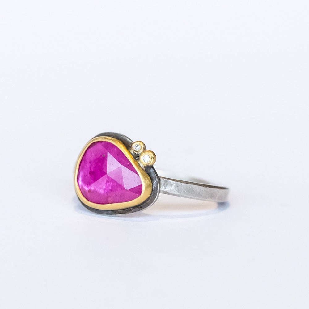 A horizontally set, teardrop shaped faceted pink ruby is bezel set in yellow gold mounted on a sterling silver ring. It is accented by two tiny bezel set diamonds.
