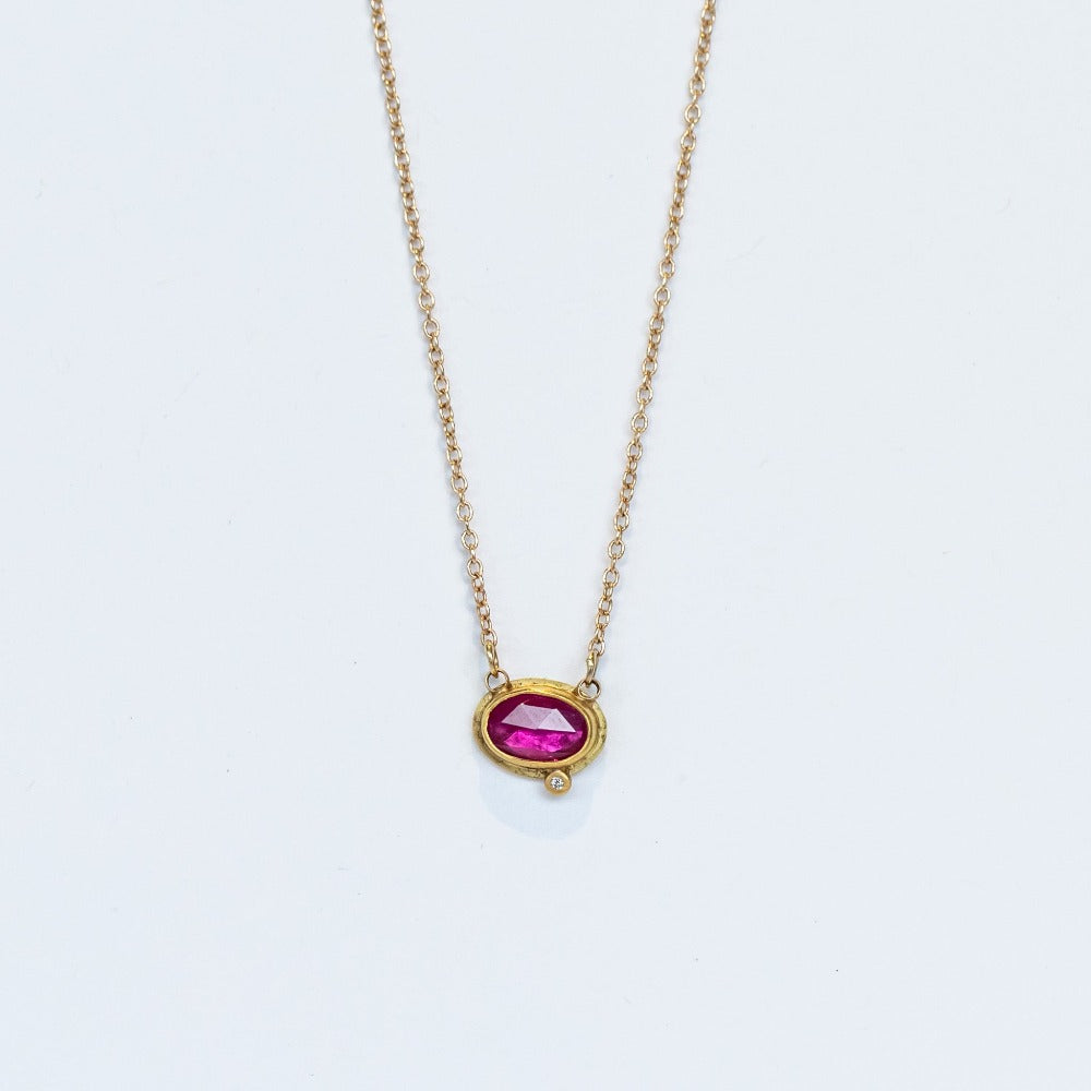 A dainty, horizontally set oval ruby is set in a yellow gold bezel with one tiny diamond accent on a gold chain.