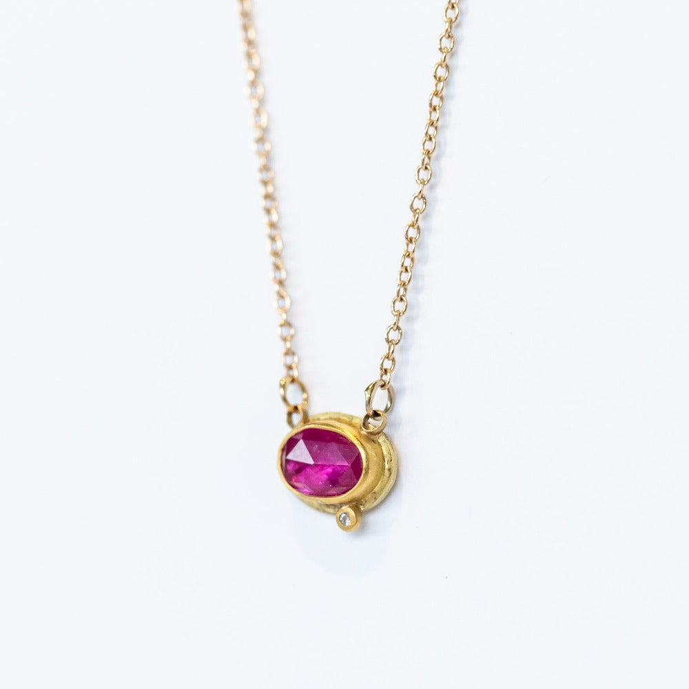 A dainty, horizontally set oval ruby is set in a yellow gold bezel with one tiny diamond accent on a gold chain.