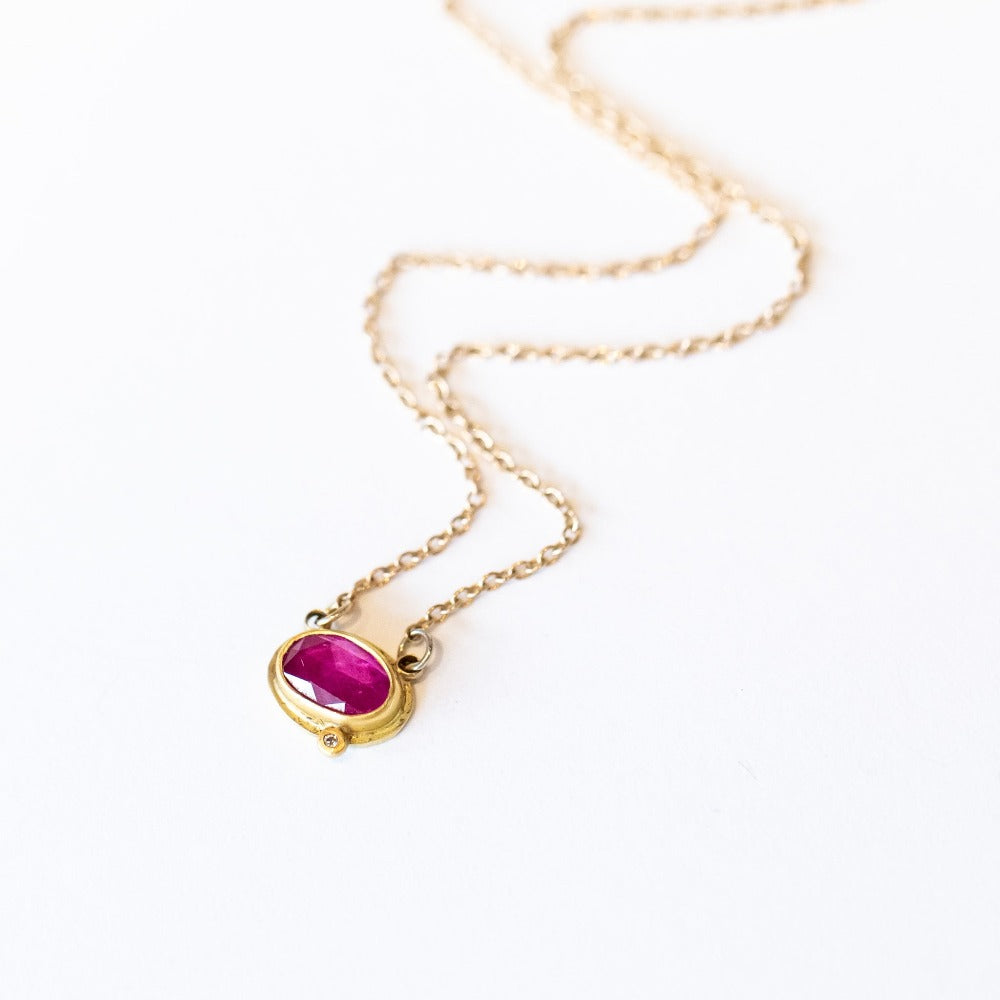 A dainty, horizontally set oval ruby is set in a yellow gold bezel with one tiny diamond accent on a gold chain.