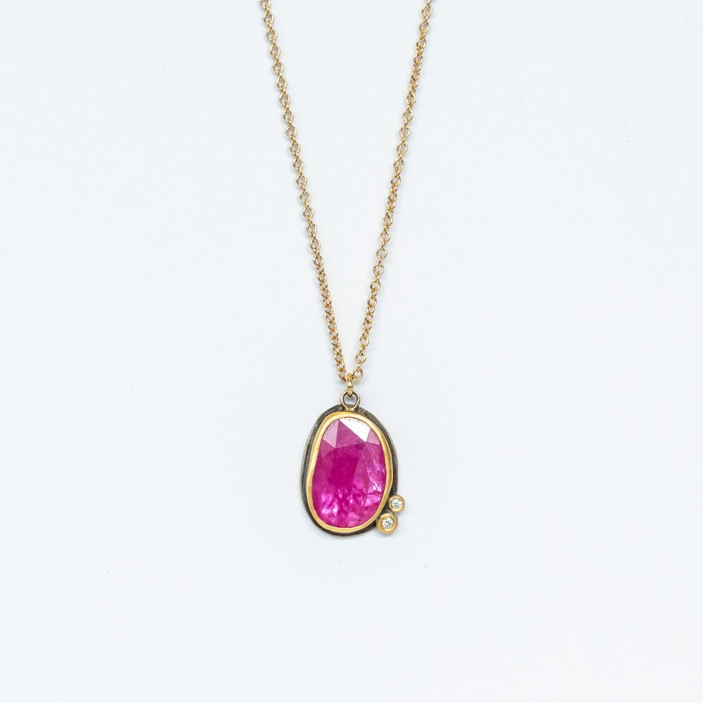 A bright pinkish-red, rose cut ruby is set with two tiny white diamond accents in 22k yellow gold bezels backed in oxidized sterling silver on a yellow gold chain necklace.