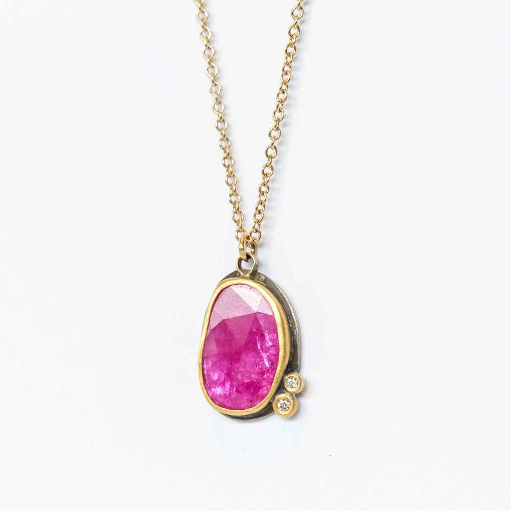 A bright pinkish-red, rose cut ruby is set with two tiny white diamond accents in 22k yellow gold bezels backed in oxidized sterling silver on a yellow gold chain necklace.